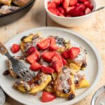 confectioner sugar dusted kaiserschmarrn on white plate with strawberry pieces scattered on top, a white bowl of strawberries and non-stick skillet with remainder of kaiserschmarrn adjacent to the plate