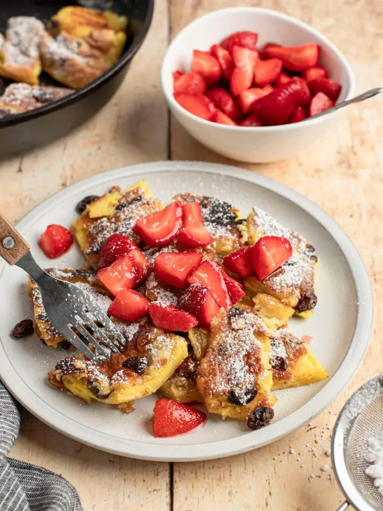 confectioner sugar dusted kaiserschmarrn on white plate with strawberry pieces scattered on top, a white bowl of strawberries and non-stick skillet with remainder of kaiserschmarrn adjacent to the plate